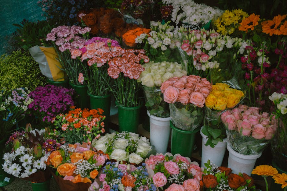 A vibrant display of fresh flowers arranged in green and white buckets and bouquets, featuring roses in shades of pink, yellow, and white; carnations in orange and white; pink and purple statice; as w