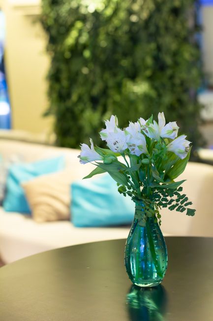 A glass vase with a textured, deep teal finish holds a fresh floral arrangement of white and pale lavender alstroemeria flowers, complemented by green fern-like foliage. The bouquet is placed on a smo