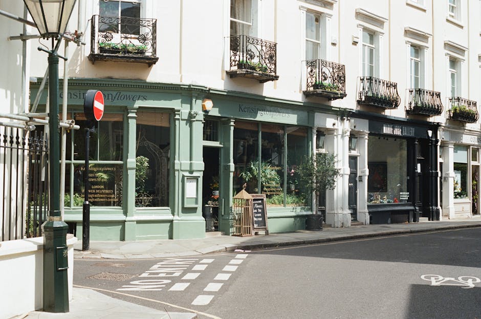 A storefront of a flower shop with a light green facade featuring large glass windows displaying various flower arrangements inside. The shop is named 'Kensington Flowers' and has a blackboard sign ou