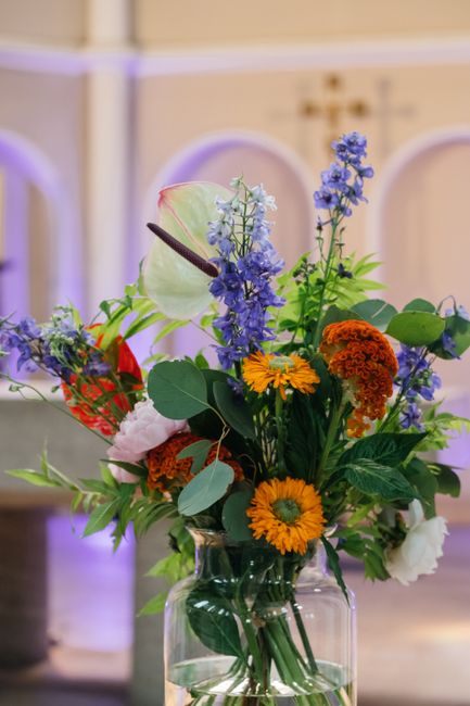 A vibrant floral arrangement in a clear glass vase featuring a variety of fresh flowers, including bright orange and yellow daisies, deep blue delphiniums, orange marigolds, pale pink roses, and lush