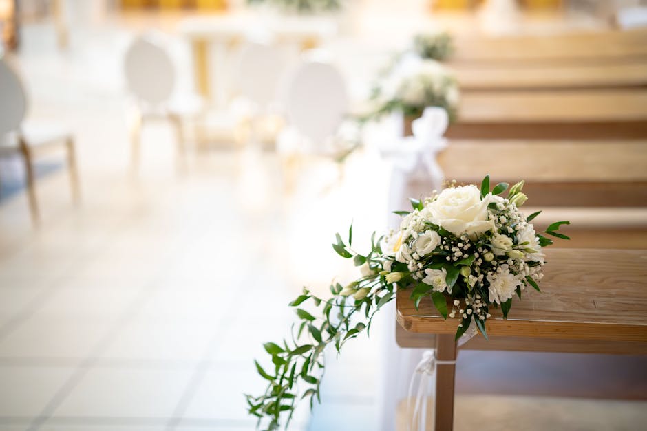 A floral bouquet featuring white roses, small white carnations, and baby's breath with green foliage, arranged in a compact, rounded style. The bouquet is placed on the edge of a wooden pew during a w