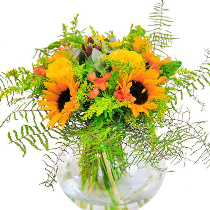 Offering a vivid celebration of warmth and light, this bouquet crafted in Waterloo is an ode to seasonal splendour and artisan skill. At a glance, the radiant yellow of towering sunflowers and plush garden roses leap forward-each bloom arranged to evoke both energy and comfort, much like the bustling markets and green pockets of Cornwall Road. Between the generous blossoms, clusters of scarlet hypericum berries glisten, reminiscent of autumn berries glimpsed in local parks. Deep, cool green foliage is woven throughout: soft, cascading fronds of fern and airy snips of light green filler interlace with bluish eucalyptus, adding depth and gentle movement to the composition. Arranged in a rounded, crystal-clear vase, the bouquet's stems become part of the visual story, refracted through the water and highlighting the freshness synonymous with Waterloo Florist. Up close, one might notice the tactile harmony-the slightly velvety texture of the sunflower petals, the silken spirals of the roses, the crispness of the greenery. Soft natural light plays across the arrangement, casting gentle shadows on the white table, enhancing the lustre of each petal. The scent is delicate yet invigorating, conjuring memories of riverside walks and balmy evenings on Lower Marsh. This striking bouquet is suited for life's brightest moments-be it a birthday, anniversary, or the start of a new chapter-reflecting the diversity and warmth of Waterloo itself.