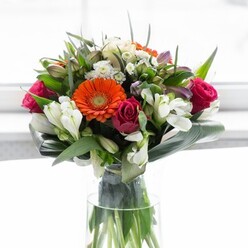 This stunning floral arrangement captures a masterful balance of colour and form, as if thoughtfully composed in a sunlit corner of Waterloo close to the National Theatre. At the heart of the bouquet, creamy peach gerbera daisies create a soft, luminous halo, their warm petals circling bold orange centers that glow with festive energy. Tucked around them are sumptuous fuchsia-magenta roses, their lush petals exuding a sense of romance and opulence, perfectly offset by the subtle presence of white alstroemeria and delicate spray roses. These white blooms infuse the design with light, while hints of pale pink and petite filler flowers offer a sense of abundance and cheerful intricacy. The entire bouquet is embraced by rich, glossy foliage-broad, paddle-shaped leaves curling gracefully around the base, their texture sturdy yet supple, reminiscent of the mature greenery of local gardens. Smaller, pointed greenery fills in every gap, adding volume and lively contrast. Displayed in a tall, clear vase, the green stems stand proud and vivid, contributing a modern, organic note to the composition. The soft, radiant background gently focuses all attention on the flowers, enhancing their hues and natural vibrancy. Ideal for sharing congratulations, expressing affection, or simply brightening a Waterloo home, this bouquet's expert craftsmanship signals a deep knowledge of both floristry and the spirit of the local community.